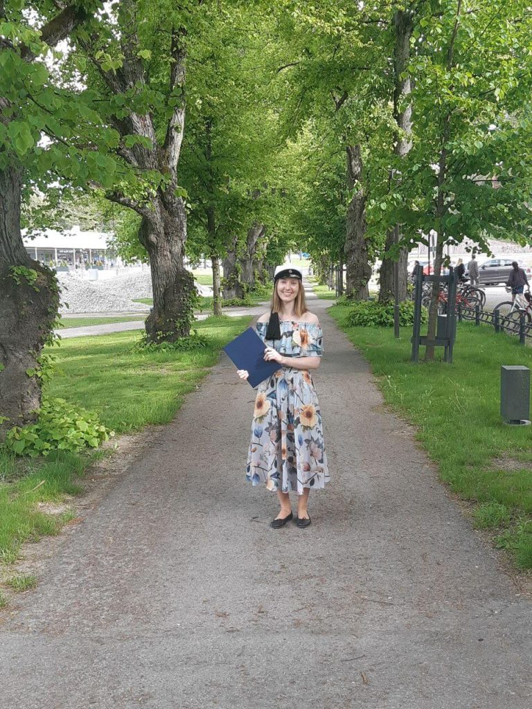 Smiling woman holding a graduation certificate while standing on a tree-lined pathway outdoors.